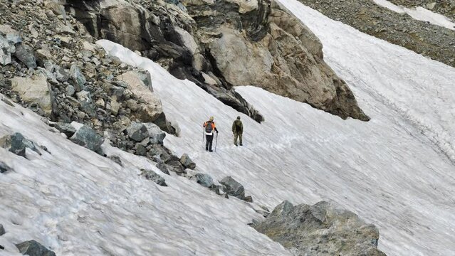 Two hikers are traversing a snowdrift in high mountains. Male travellers moving along a path trampled in the snow. Summer hike in the Caucasus mountains