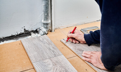 Close up of man construction worker with pencil in hand drawing mark on laminate wooden plank. Male worker preparing laminate material for floor installation in apartment under renovation.