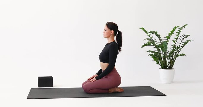 Brunette woman performs extension and flexion of the spine in vajrasana, trains in sportswear while sitting on a mat in the room