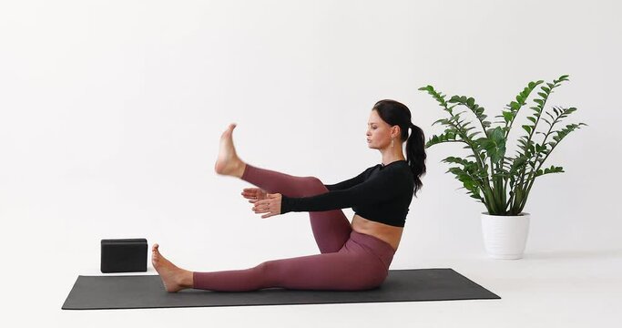 Yoga woman performs Paripurna Navasana exercise, boat pose with alternating leg lifts and foot warm-up, training in leggings and a short long-sleeved top while sitting on a mat