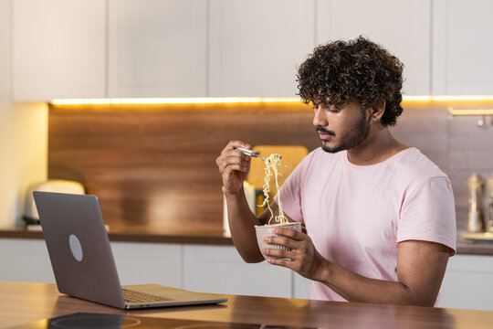 A mestizo young man stands sideways in the kitchen in subdued lighting, holds a bowl of pasta in hands and wraps them in a fork. An american or african guy student uses a laptop, enjoys a dinner and