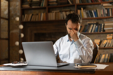 A man squints eyes and covers face with hand tiredly, sits at a table in the library and uses a...