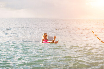 Woman works on laptop in sea. Freelancer, blond woman in sunglases floating on an inflatable big pink donut with a laptop in the sea at sunset. People summer vacation rest lifestyle concept.