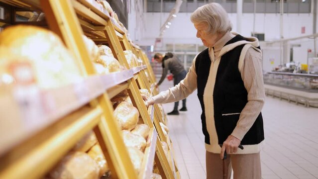 An old woman with gray hair chooses bread in a large supermarket, she stands near the shelves with bread and reads the composition in the products.