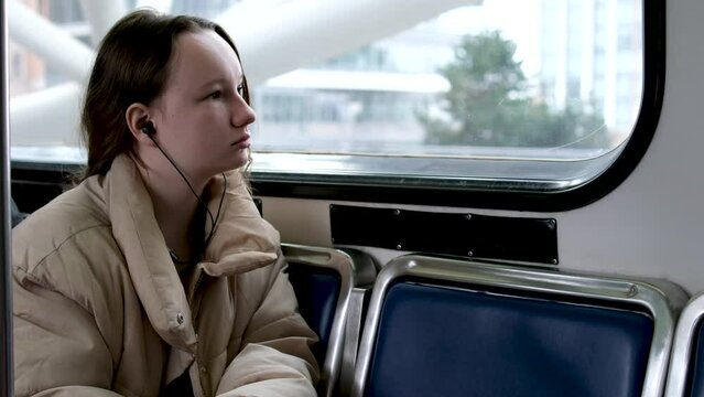teenage girl rides in skytrain on train in bus headphones in ears listens to music autumn spring winter warm beige jacket hair is tied down real person sits tucked legs under sad dreams awaits stop