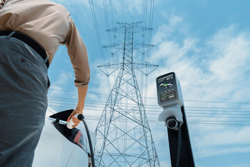 Man recharge EV electric car battery at charging station connected to electrical power grid tower...
