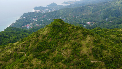 Mountain range of a tropical island. Mountain tops covered with jungle. Aerial view of green hills, mountain peaks, paths on the slope and the town behind.