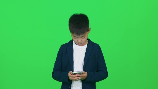 Focused asian schoolboy in a suit standing on a green background and using a smartphone, template on a chromakey.