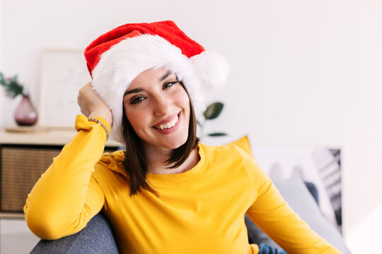 Friendly portrait of young millennial female in Santa Claus hat smiling at camera relaxing on sofa at christmas vacation at home