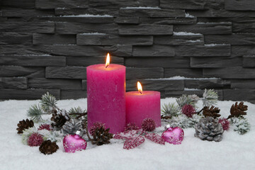 Pink Christmas candles with branches, pine cones on a blanket of snow in front of a gray brick wall...