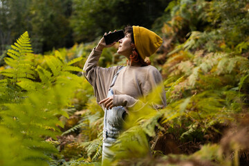 Woman with mobile phone and headphones in fern forest wearing yellow hat, listening to music, taking photos and video call