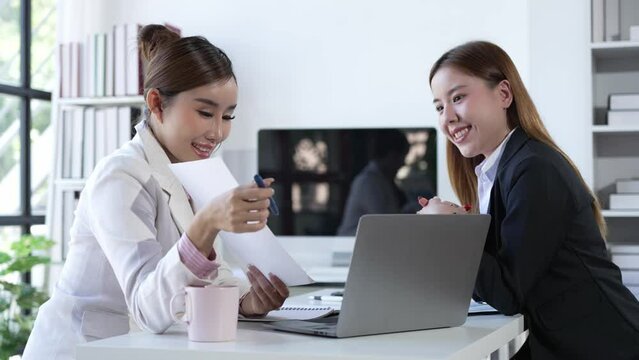 Two Asian businesswomen working on laptop computers discuss financial data in marketing project document analysis. 4k