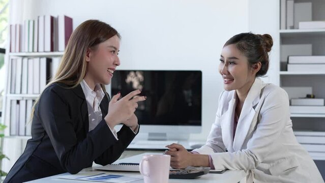 Two Asian businesswomen working on laptop computers discuss financial data in marketing project document analysis. 4k