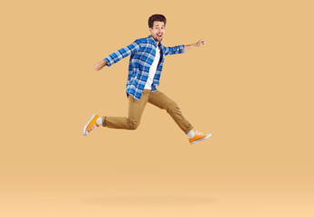 Fototapeta na wymiar Handsome happy guy jumping high in air on beige background with copy space. Full length photo of positive cheerful hurrying young man wearing casual plaid shirt trousers running away fast