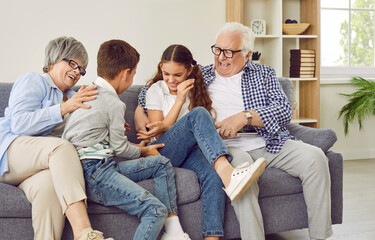 Happy grandparents and children having fun on the weekend. Funny, cheerful grandmother and grandfather tickling happy little kids grandchildren while sitting on the sofa at home all together - obrazy, fototapety, plakaty
