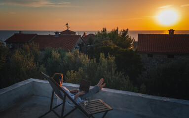 Happy young woman is working using laptop sitting on the patio in summer at sunset