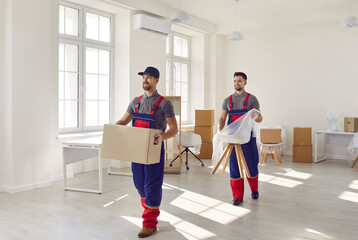 Team of two young cheerful professional workers of moving service in overalls carrying and...