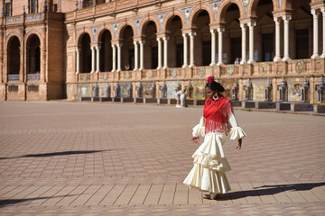 Fototapeta na wymiar Young black and South American woman in a beige gypsy flamenco suit and red shawl, posing in a beautiful square in the city of Seville in Spain. Concept dance, folklore, flamenco, art.
