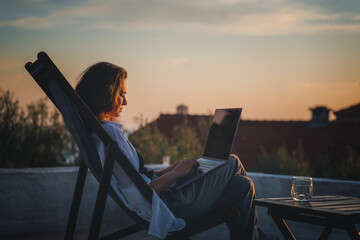 Happy young woman is working using laptop sitting on the patio in summer at sunset - obrazy, fototapety, plakaty