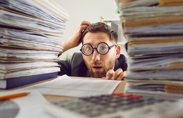 Bookkeeper is shocked at the load of work. Portrait of a young man in round glasses between two stacks of paperwork on the office table looking at the camera with a funny surprised facial expression - obrazy, fototapety, plakaty