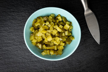 Small bowl filled with diced pickles placed on black background next to small kitchen knife