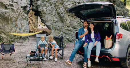 Parents father and mother sitting in car trunk at camping and child resting in shade of tree near the car in nature have picnic break for relaxing next trip before forest hike. Outbound family trip