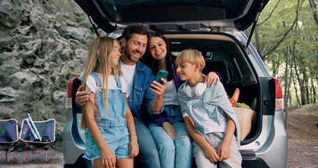 Cheerful lovely parents with cute teen kids which using mobile phone sitting in the car's trunk....