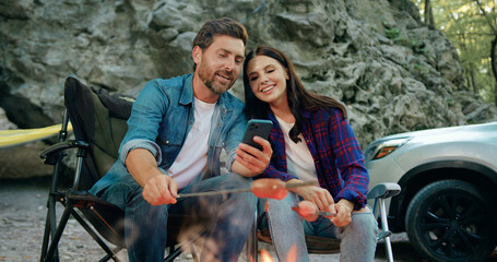 Happy family couple man and woman using phone sitting in forest near car while traveling during summer vacation road trip. Pretty family mother father roasting sausages by bonfire talking and smiling
