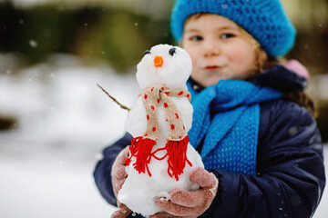 Cute little toddler girl making mini snowman and eating carrot nose. Adorable healthy happy child playing and having fun with snow, outdoors on cold day. Active leisure with children in winter