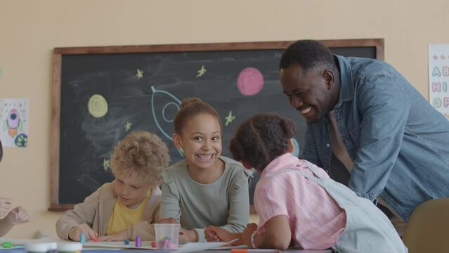 African American teacher smiling and talking with multi-ethnic kids drawing picture together with crayons during art class in primary school