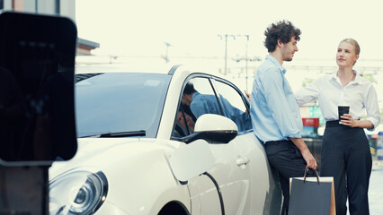 Progressive businessman and businesswoman leaning on electric car connected to charging station...