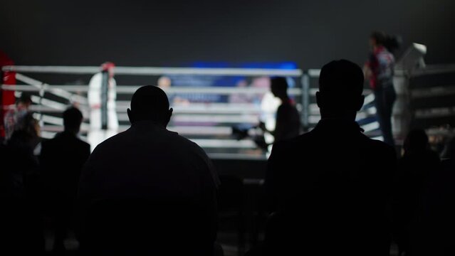 Men watch the fight of karatekas in the ring. Silhouettes of people, general plan.