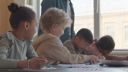 Cheerful multi-ethnic kids having fun while drawing with crayons during art class in primary school, African American teacher walking behind and helping