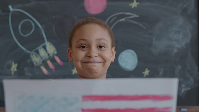 Little African American girl holding paper drawing of US flag in front of camera, then revealing her face and posing with smile in school classroom