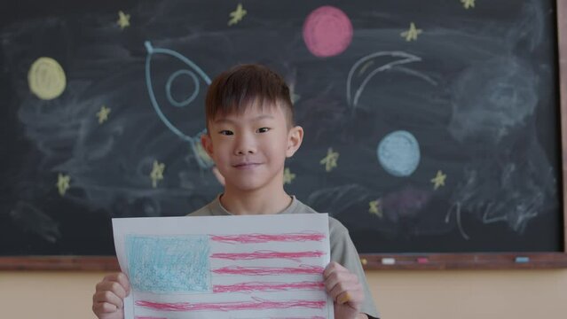 Portrait of little Asian boy holding paper drawing of American flag and posing for camera with smile against chalkboard with space rocket drawing in school classroom