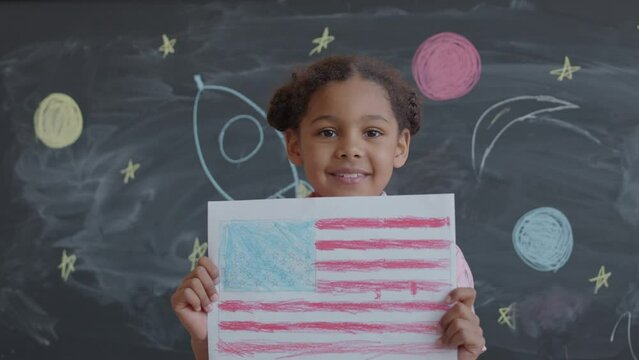 Portrait of little African American girl holding US flag paper drawing and smiling on camera while standing by chalkboard with space rocket drawing in school classroom