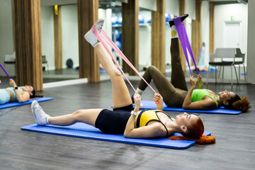 A group of 3 women of different ethnicities are doing a Pilates class using elastic bands inside a...