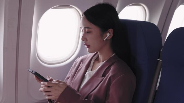 Asian businesswoman is actively using her phone to record and analyze work while wearing headphones during her flight. She concentrates on her tasks, utilizing the flight time efficiently.