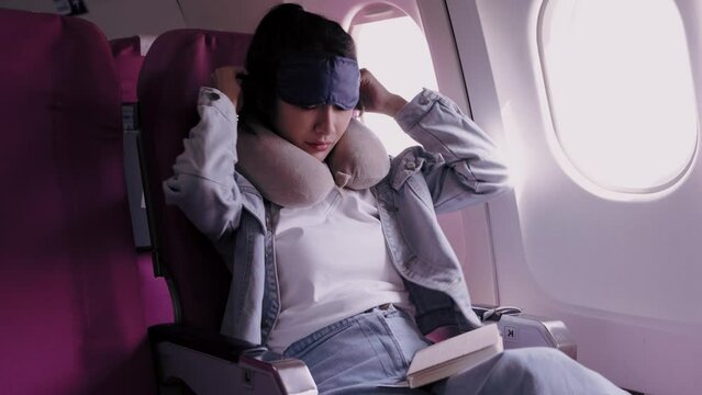 Asian female tourist reads a book, covers her eyes to avoid light, and takes short naps during her journey to a holiday destination on her days off. She travels by plane for convenience.