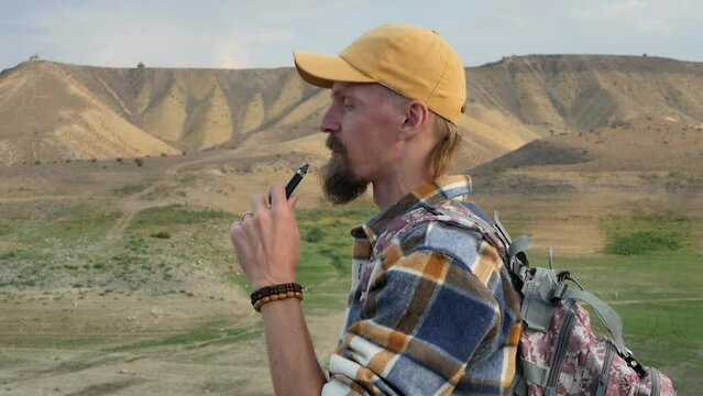 A Caucasian young tourist with a beard and a yellow cap smokes an electronic cigarette of the pod system while standing against the backdrop of beautiful sandy mountains and exhaling steam upward.