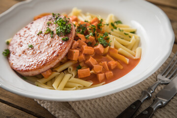 Jägerschnitzel nach DDR GDR Rezept mit Jagdwurst gebraten, Makkaroni Penne Nudeln, Tomatensoße, Petersilie mit Porzellan tiefer Teller, Besteck, Salzstreuer, Serviette, rustikal dunkel Holz Hintergund