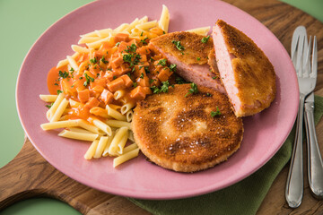 Jägerschnitzel nach DDR GDR Rezept mit Jagdwurst paniert, Makkaroni Penne Nudeln, Tomatensoße, Petersilie mit pink rosa Teller, Besteck, Holzbrett, Serviette und grünen Hintergrund