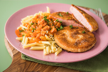 Jägerschnitzel nach DDR GDR Rezept mit Jagdwurst paniert, Makkaroni Penne Nudeln, Tomatensoße, Petersilie mit pink rosa Teller, Besteck, Holzbrett, Serviette und grünen Hintergrund