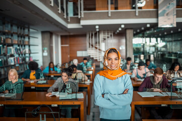 Portrait of a smiling young Arabian woman in hijab standing in library. Young female student in busy library looking at camera with confidence