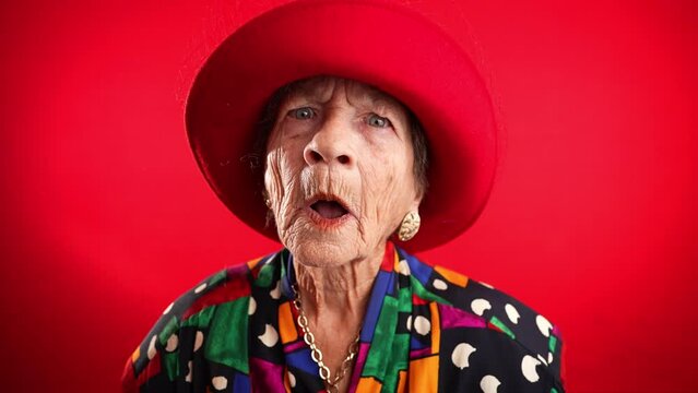 Saying WOW, a happy fisheye portrait caricature of funny elderly woman with red hat isolated on red background.