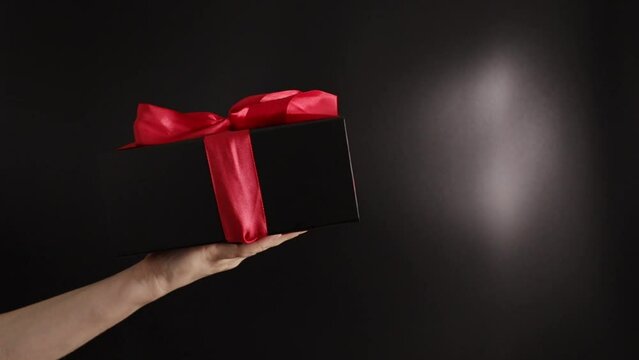 Hand with a black present box with red ribbon, elegant surprise, Festive gift on a dark background