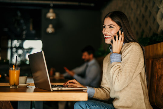 Smiling Latina woman using a smartphone and working on a laptop in the bright cafe. Man in the background. - Powered by Adobe