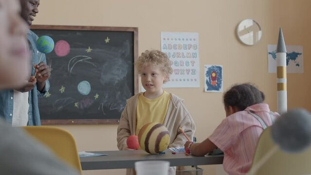 Little Caucasian boy standing in front of classmates and answering questions of African American teacher during astronomy lesson