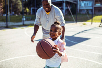 Adorable little girl playing basketball with father on outdoor court