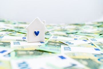A little white house with a blue heart over many euro money bills, concept of earning for a home - obrazy, fototapety, plakaty